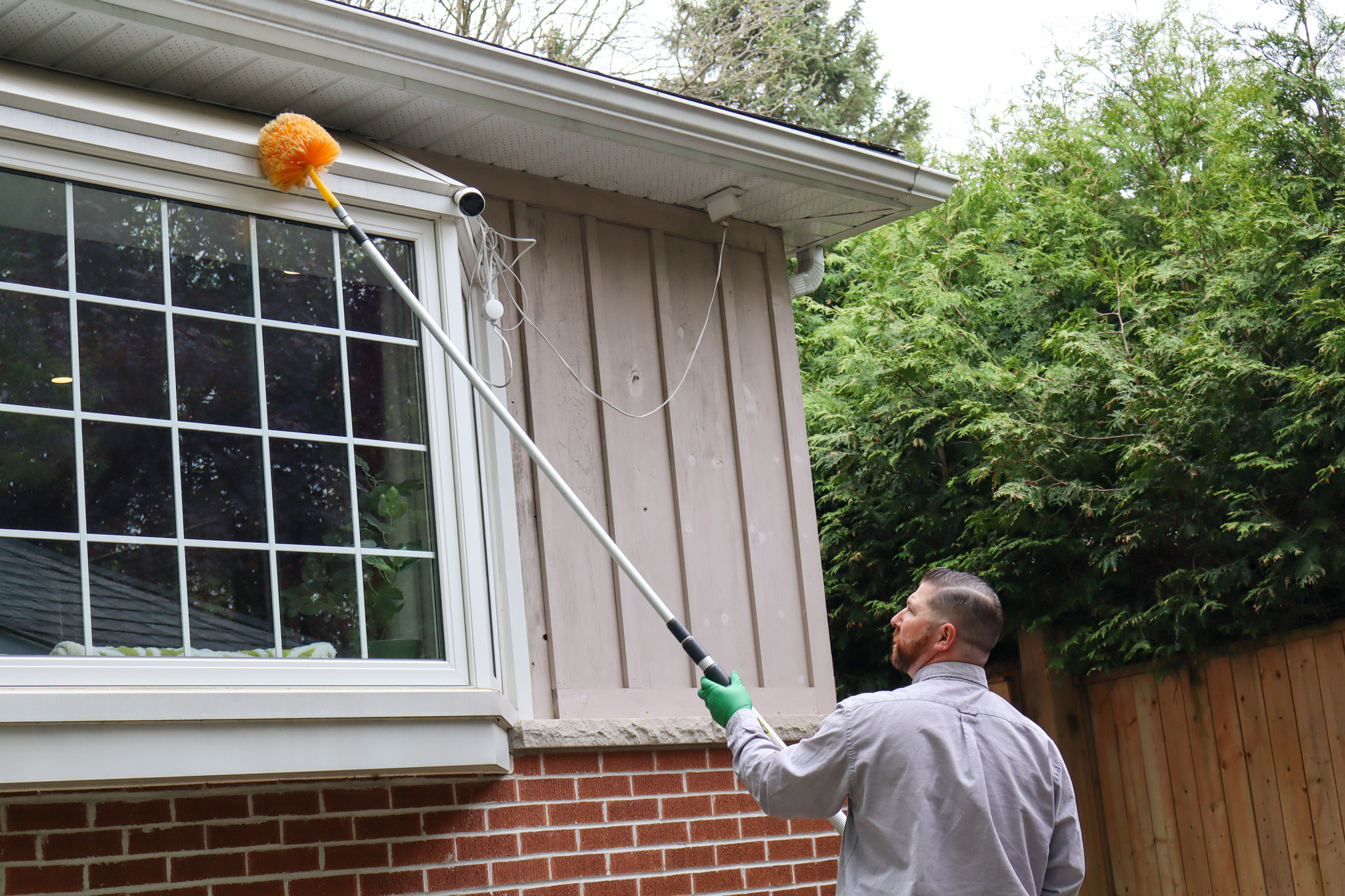 A Skedaddle technician using an extended telescopic pole with a yellow cobweb brush to clean the upper trim of a large residential bay window.