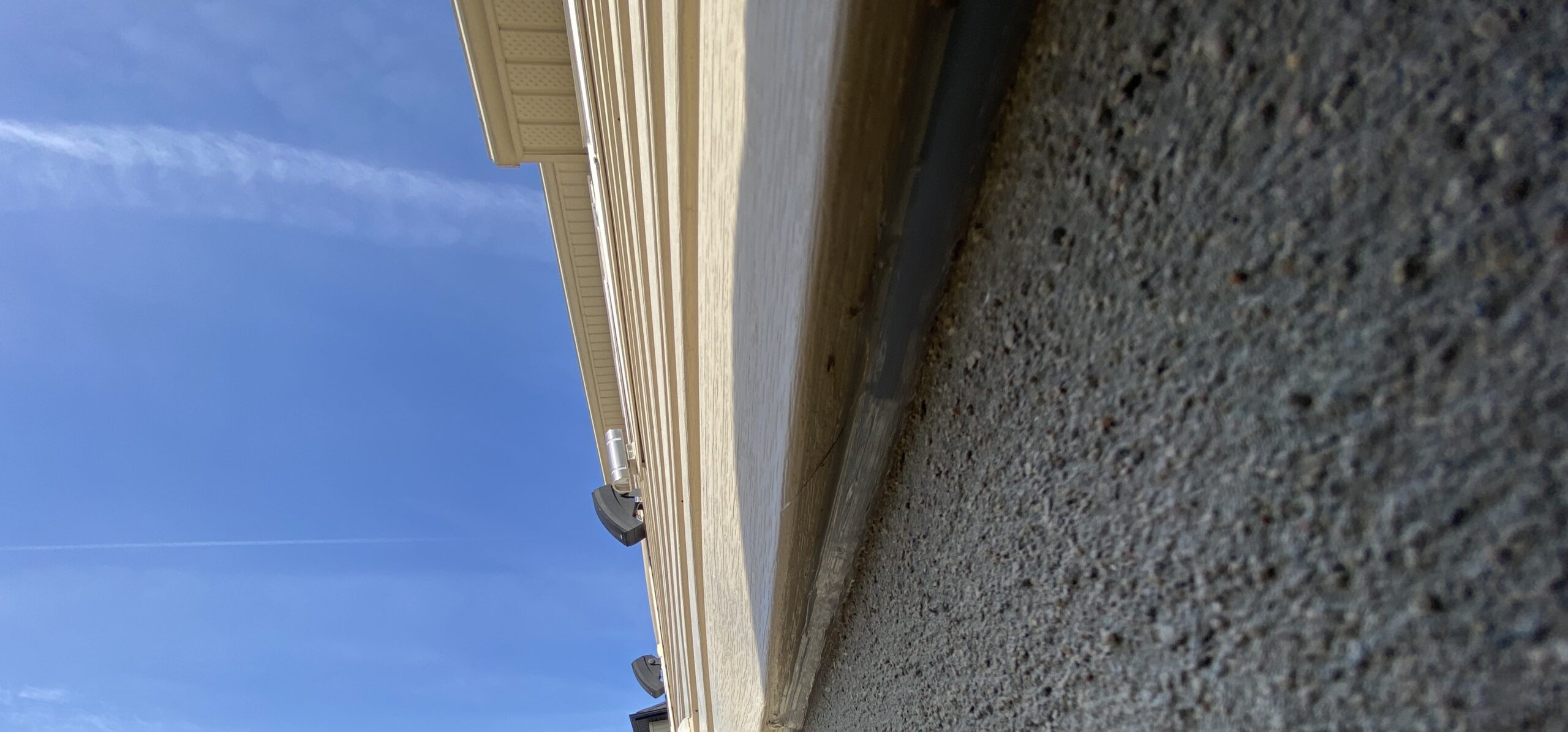 A close-up, low-angle shot looking up along the side of a house where cream-colored siding meets a grey concrete foundation, showing potential entry gaps for pests.