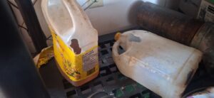 Two plastic jugs on a garage shelf, including a yellow canola oil bottle, showing large holes chewed through the plastic by destructive rodents.