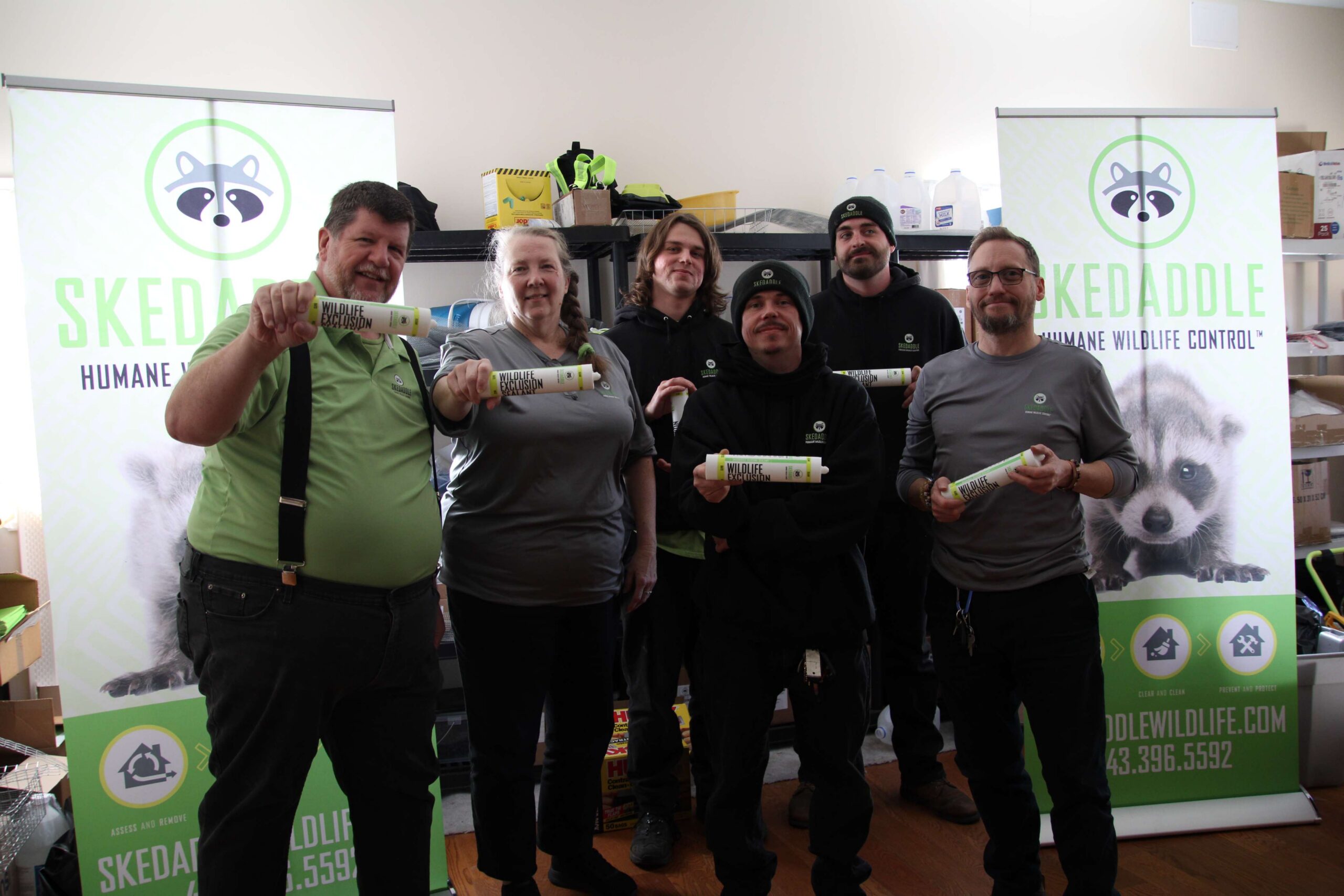 A group of Skedaddle wildlife technicians standing together and holding tubes of specialized wildlife exclusion sealant in front of company banners.