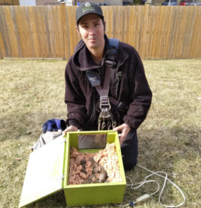 A Skedaddle Wildlife technician kneeling on a lawn holding a bright green heated reunion box containing baby raccoons nestled in insulation for safe mother-offspring reunification.