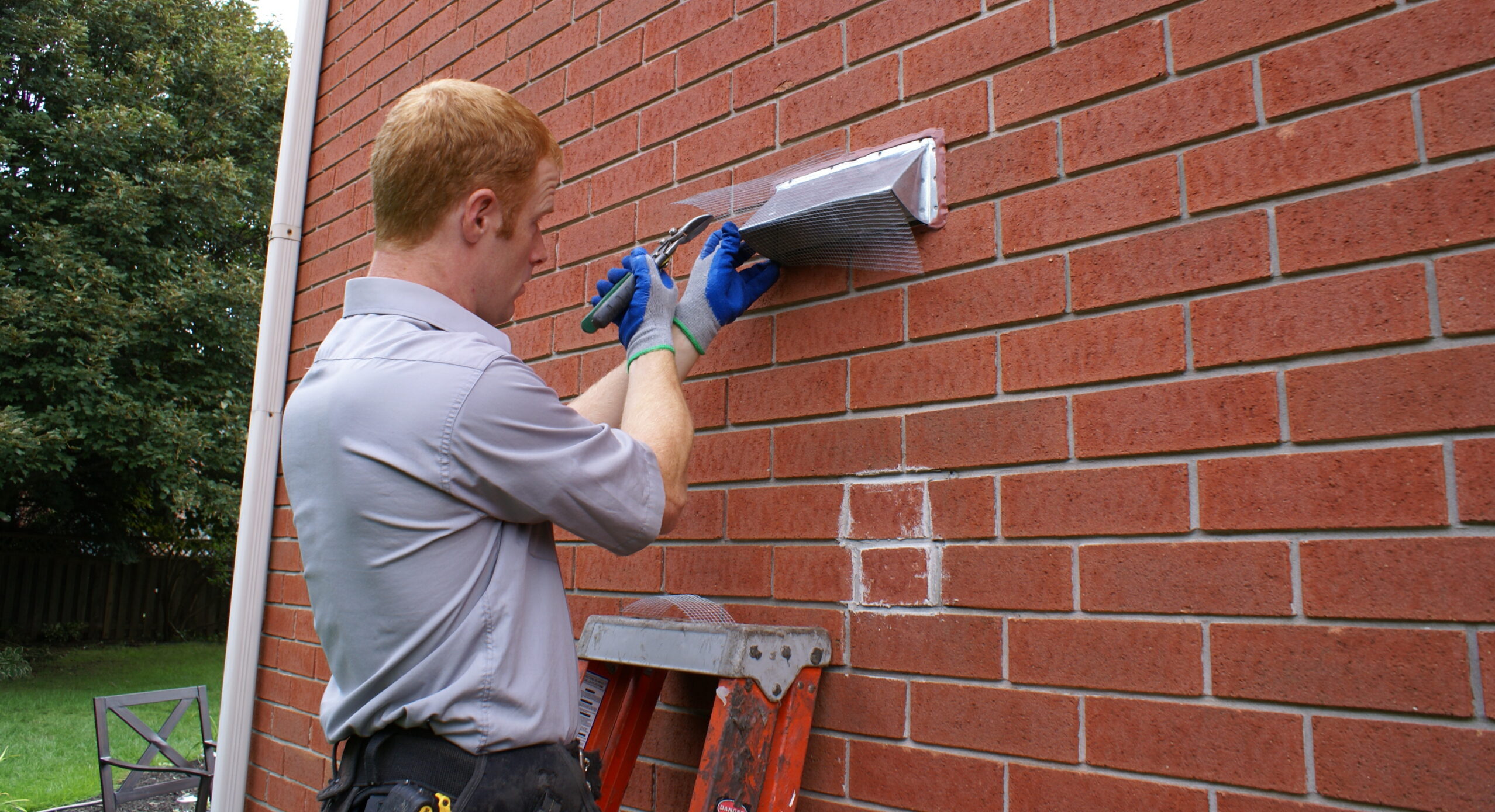Skedaddle technician in a grey shirt and blue gloves using wire cutters to install protective mesh screening over a wall vent on a red brick house in Richmond Hill.