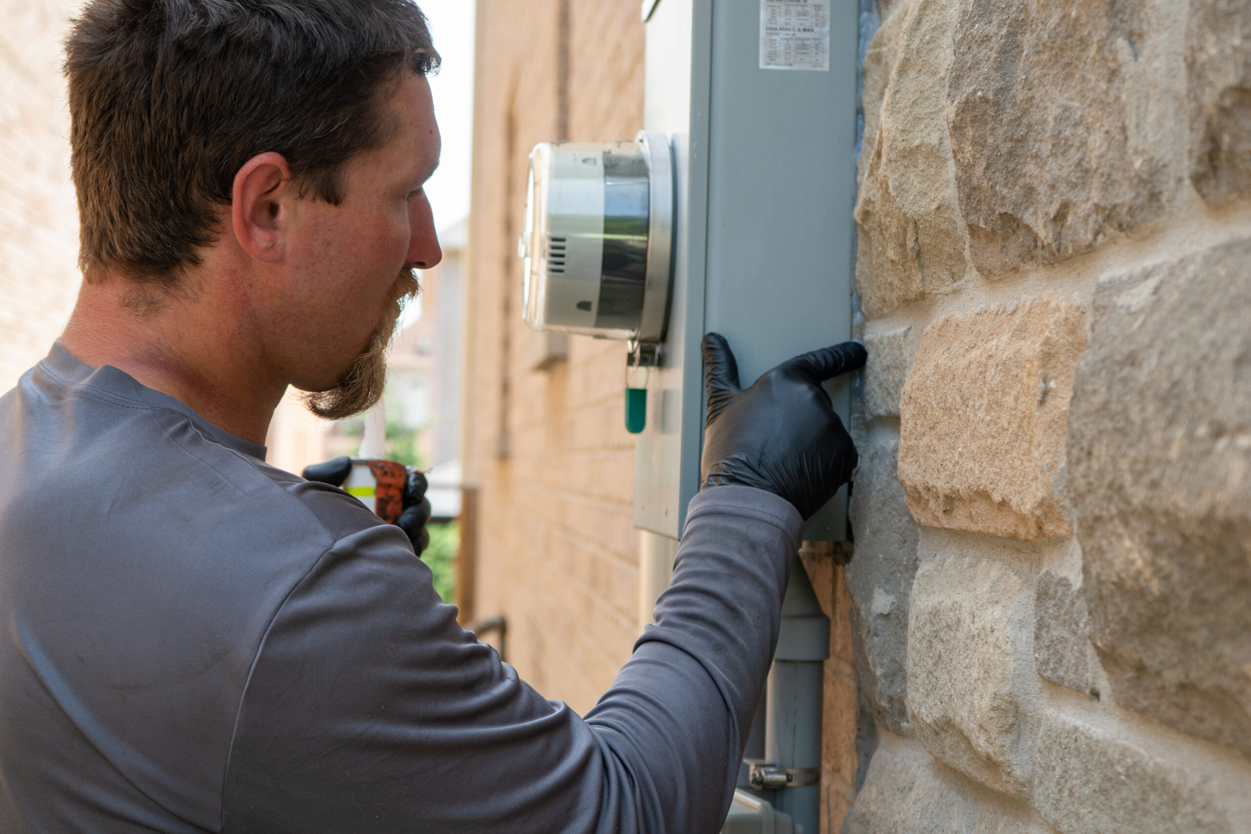 Skedaddle technician wearing black gloves sealing a potential wildlife entry point behind an electrical meter on a stone house.
