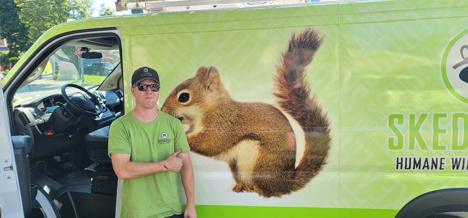 A smiling Skedaddle Humane Wildlife Control technician wearing a branded green t-shirt and hat, standing beside a lime-green service truck with a large squirrel graphic.