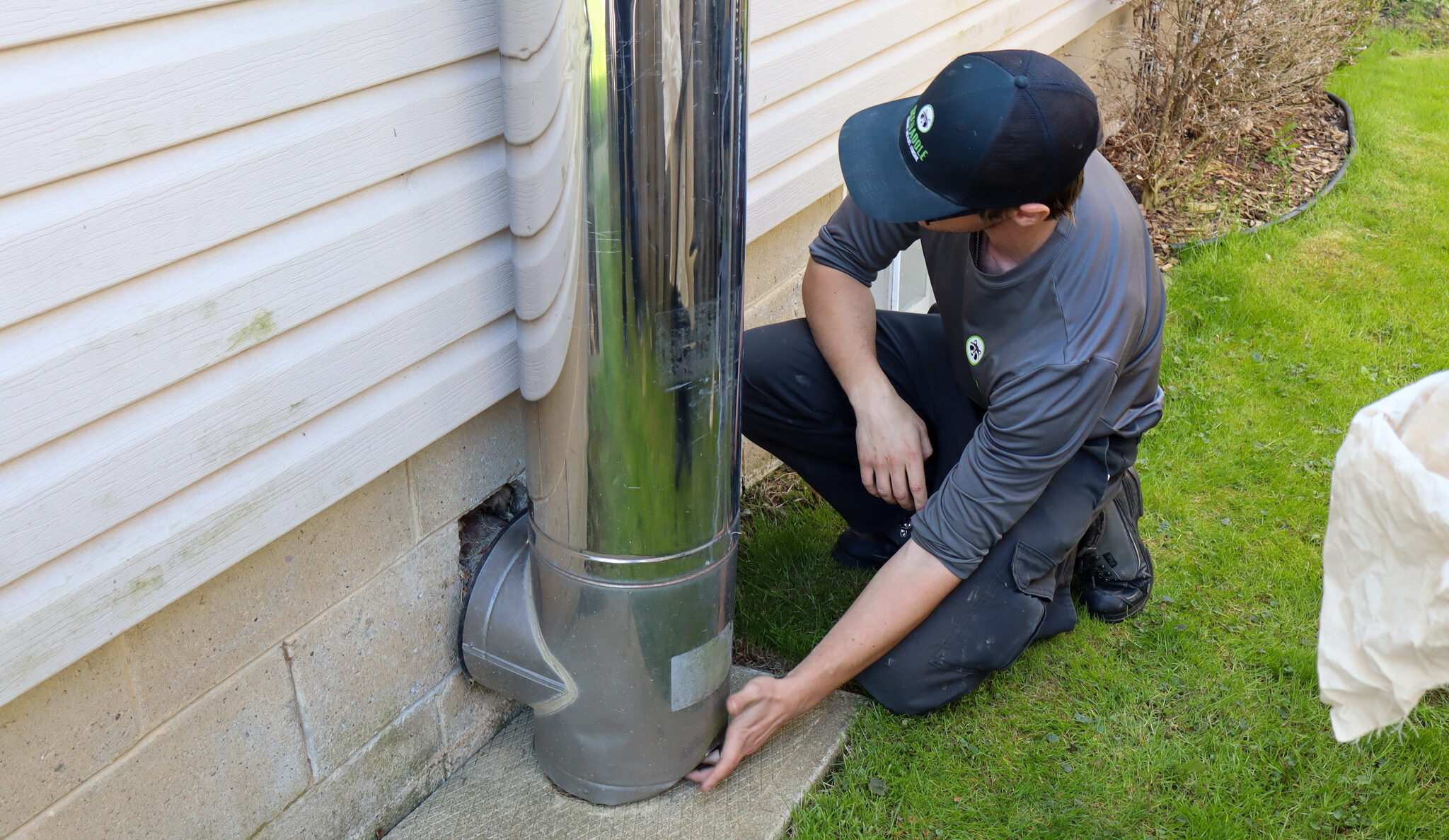 A Skedaddle wildlife technician crouching on a lawn to inspect the base of a large metal chimney exhaust pipe connected to a house foundation for potential animal entry holes.