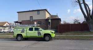 A bright green Skedaddle Humane Wildlife Control truck parked on a residential street with a technician on an extension ladder inspecting a roof