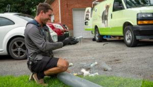 A wildlife control technician kneels on a driveway holding a one-way door device while preparing wire exclusion mesh, with a service van behind him.