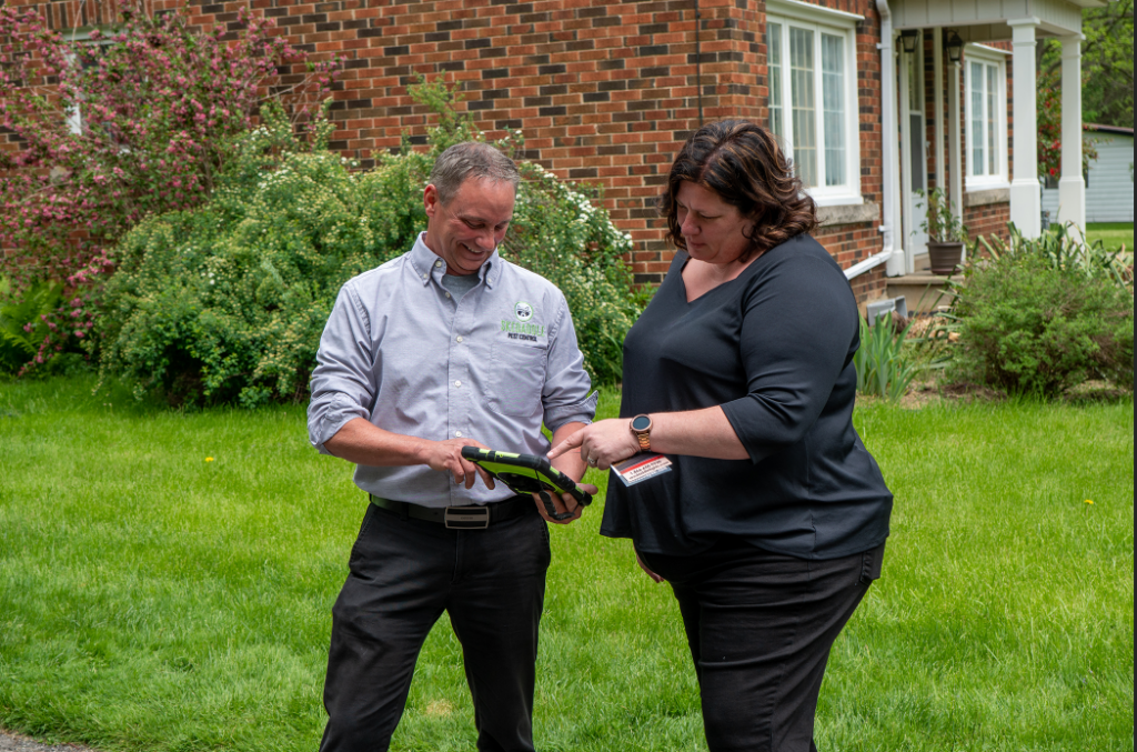 Skedaddle Pest Control Representative Reviewing Service Details on Tablet with Homeowner A Skedaddle Pest Control specialist in uniform sharing digital assessment findings on a tablet with a homeowner in her front lawn