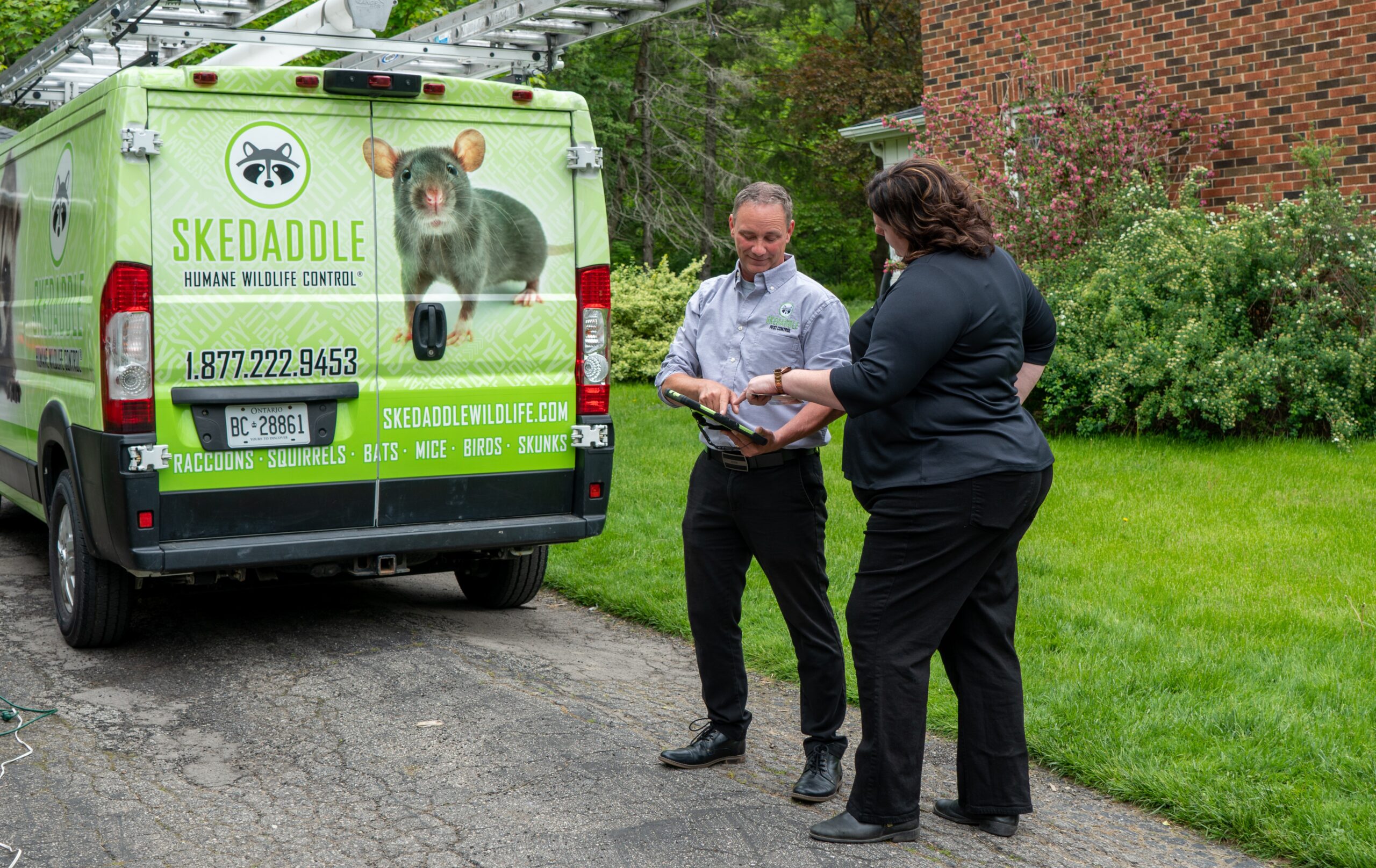 A Skedaddle Humane Wildlife Control technician showing an inspection report on a tablet to a female homeowner beside a branded work van
