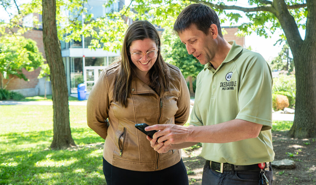 A Skedaddle technician wearing a branded polo shirt showing photos on a smartphone to a smiling female customer outdoors