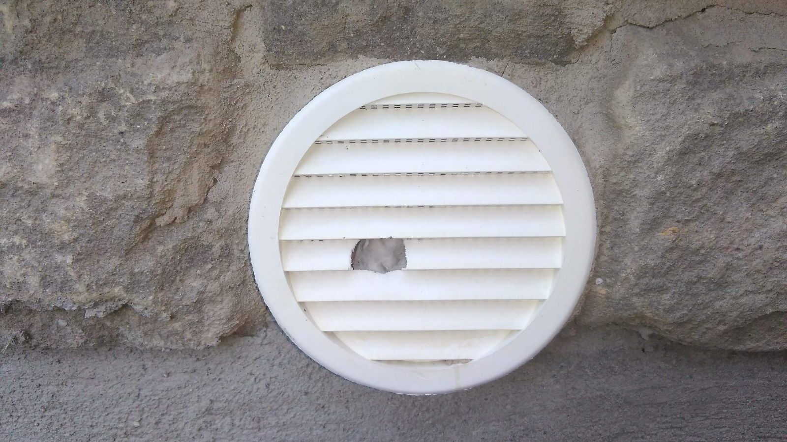 A white circular louvered vent cover mounted on a stone wall with a hole chewed through the slats