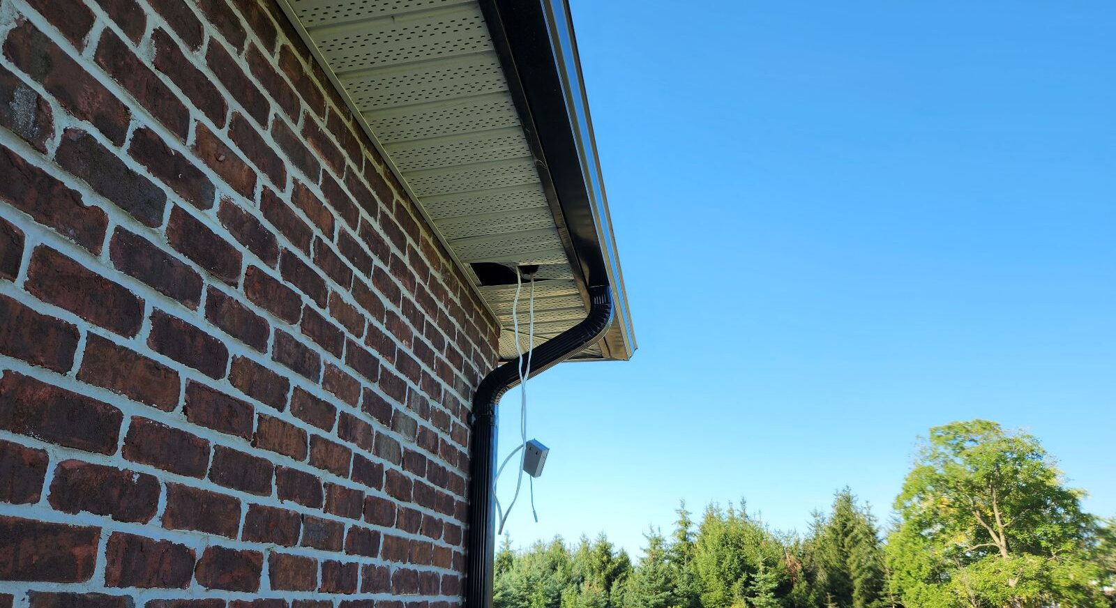 A hole in the perforated white vinyl roof soffit along a brick house with dangling electrical wires creating a potential wildlife entry point