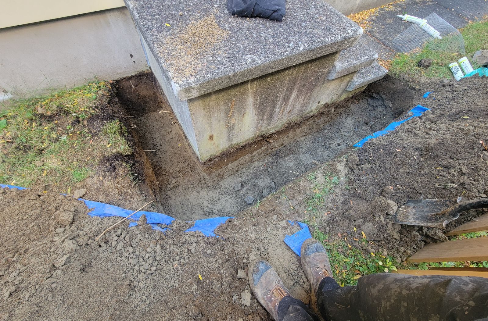A deep trench dug into the soil around the base of concrete porch stairs prepared for underground wire mesh exclusion installation