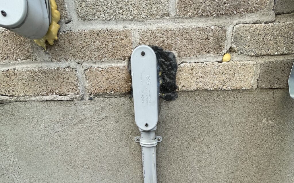 Exterior Electrical Conduit Penetration Sealed Against Mouse Entry on Concrete Wall An exterior vertical electrical conduit pipe entering a concrete and brick wall sealed with dark sealant around the opening