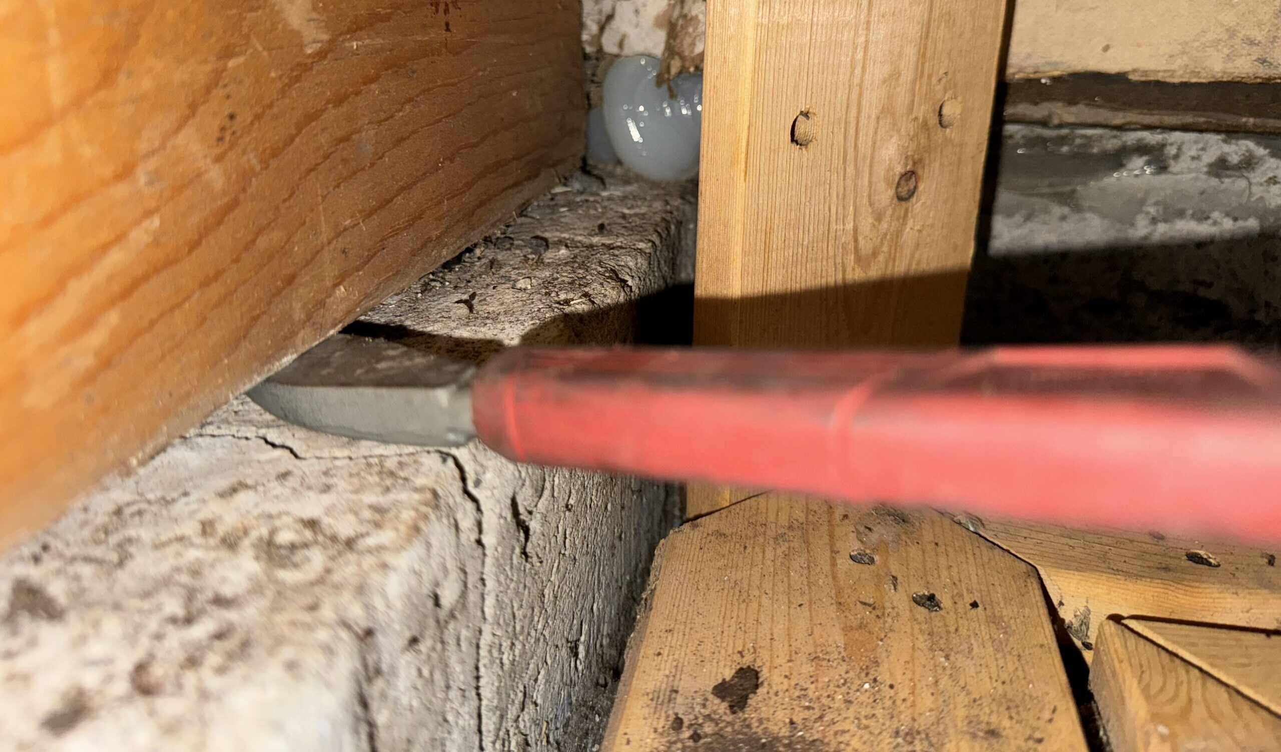 A red-handled pry bar wedged under a wooden sill plate on a concrete foundation during an inspection for mouse entry voids